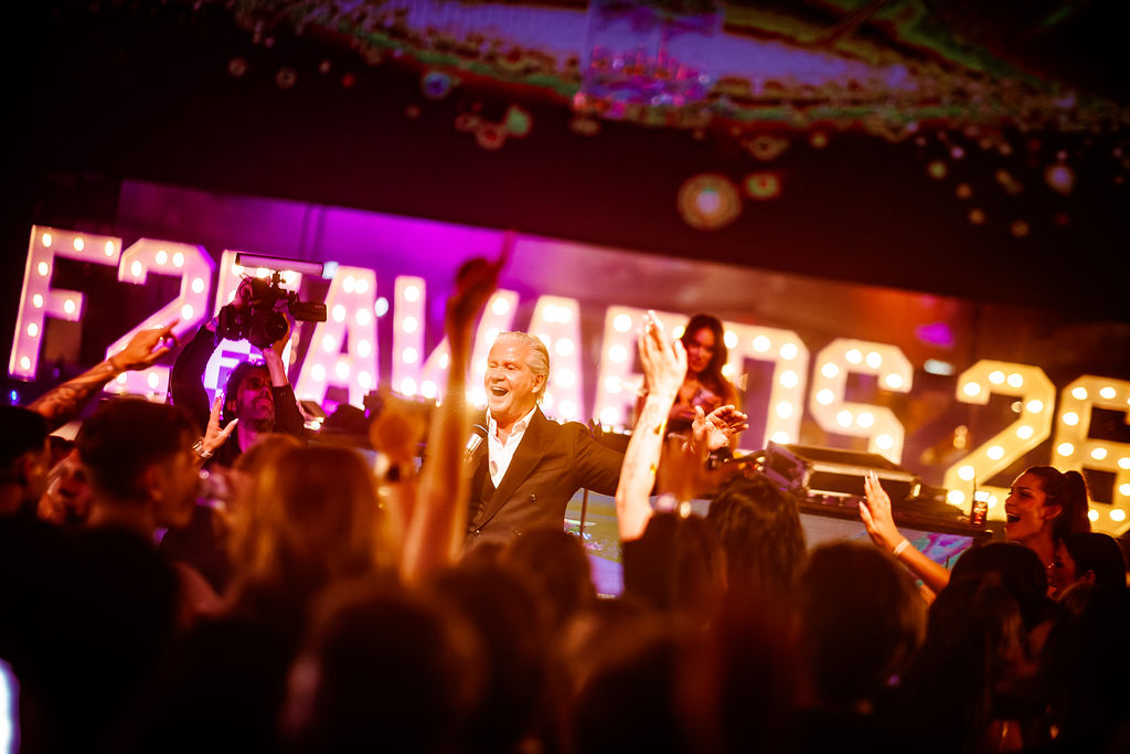 The afterparty of an award show: the host on stage with his arms wide, guests cheering, lit letters spelling the awards name behind him