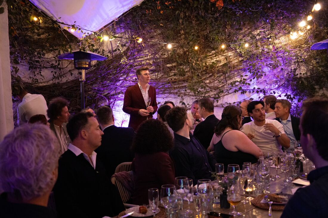 Guests seated at a long dinner table under vines listen to the host, who stands and speaks in the purple light