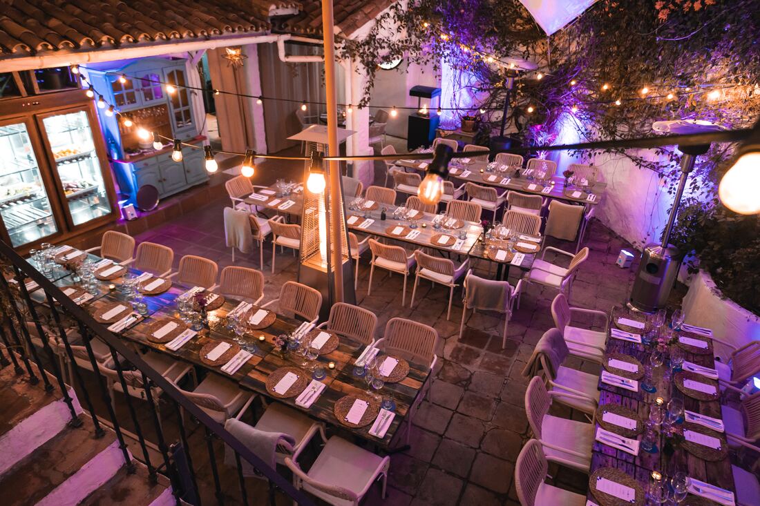 Long dinner tables set under vines and string lights in a courtyard, lit in purple, before the guests arrive