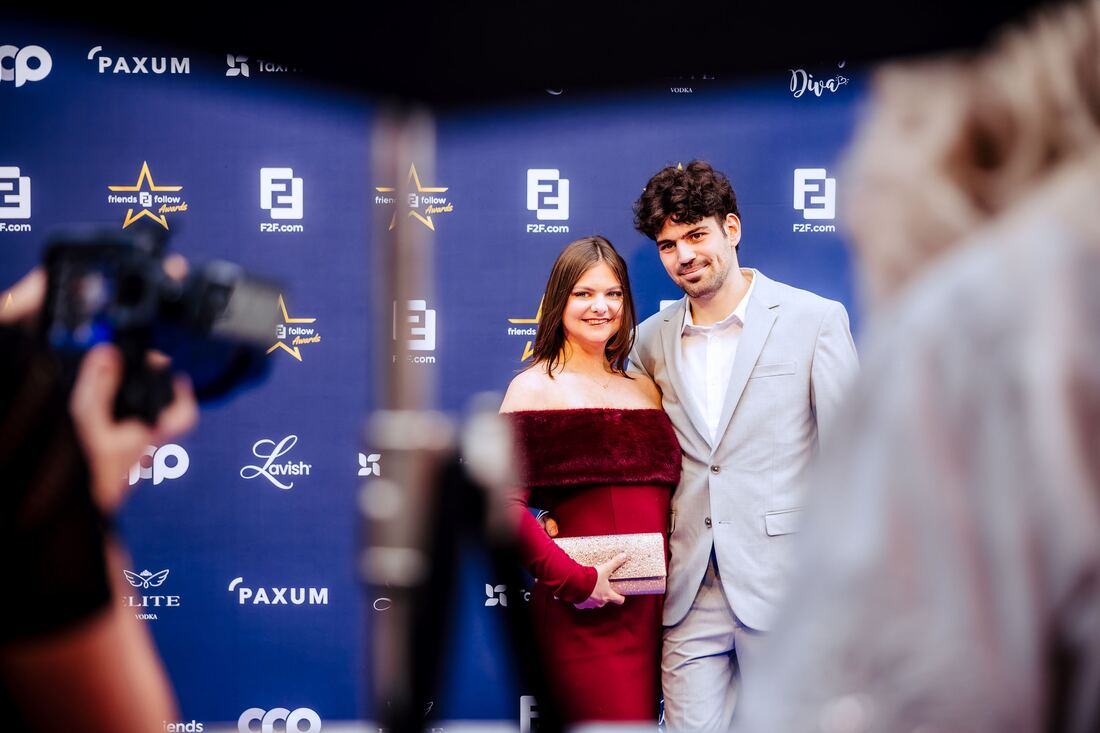 A couple in evening wear poses for photographers in front of the blue press wall with sponsor logos at the F2F Creator Awards 2026
