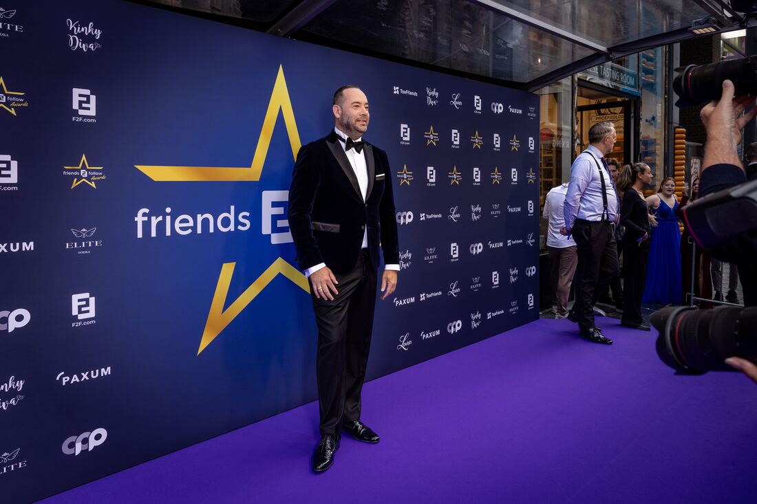 A guest in a velvet dinner jacket on the purple carpet in front of a sponsor wall, photographers on the right